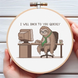 Puede incluir: Un bordado de punto de cruz en un bastidor de madera redondo que muestra un perezoso marrón con una corbata verde, sentado en un escritorio con una computadora. El texto sobre el perezoso dice I WILL BACK TO YOU QUICKLY. Una taza pequeña está.