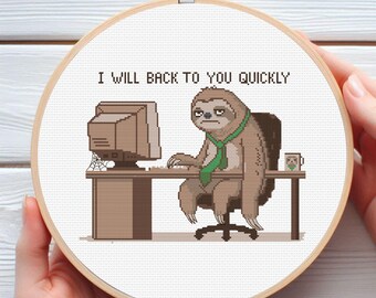 May include: A cross-stitch embroidery in a round wooden hoop featuring a brown sloth wearing a green tie, sitting at a desk with a computer. The text above the sloth reads I WILL BACK TO YOU QUICKLY. A small mug sits on the desk next to the sloth.
