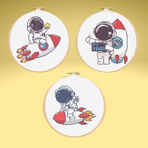 May include: Three round embroidery hoops with cross-stitch designs of astronauts. One astronaut is on a rocket, another holds a sign that says "TO MARS", and the third is near a rocket. The designs are in white, red, and yellow.