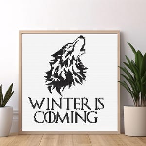 May include: A framed cross-stitch featuring a black howling wolf silhouette above the text "WINTER IS COMING" in a bold, serif font. The artwork is set against a white background, with a wooden frame. Decorative plants flank the framed art.
