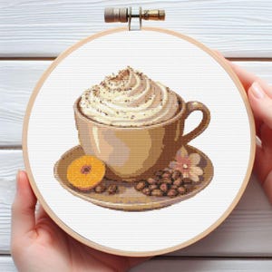 May include: Cross-stitch embroidery of a coffee cup with whipped cream, coffee beans, and a slice of orange on a saucer. The embroidery is set in a wooden hoop, held by hands. The cup and saucer are shades of brown and beige.
