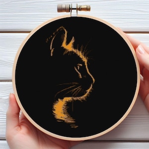 Puede incluir: Un bordado de punto de cruz de un perfil de gato en un aro de madera. La silueta del gato es en tonos naranja sobre un fondo negro. El aro de bordado está sostenido por dos manos.