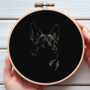 May include: A black and white cross-stitch embroidery of a German Shepherd dog's head, set in a wooden embroidery hoop. The dog's face is detailed with visible fur texture against a solid black background. The hoop is held by hands.