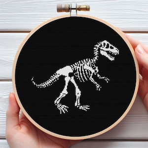 Può includere: Un disegno a punto croce di uno scheletro di dinosauro bianco su sfondo nero, esposto in un telaio da ricamo in legno. Il dinosauro è un Tyrannosaurus Rex. Il telaio è tenuto da una persona.