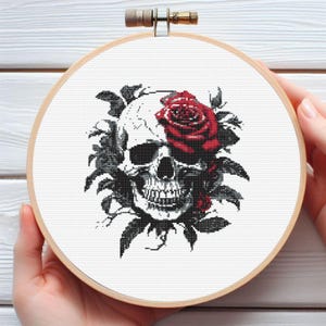 May include: Cross-stitch embroidery in a wooden hoop featuring a skull with a red rose and dark leaves. The skull is detailed in black and white, with the rose adding a pop of color. The design is on a white background.