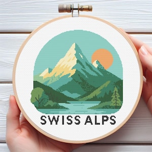 Puede incluir: Un bastidor de bordado de punto de cruz con un diseño de paisaje de los Alpes suizos. El diseño incluye una cordillera, un lago, árboles y un sol poniente. El texto "SWISS ALPS" está en la parte inferior.
