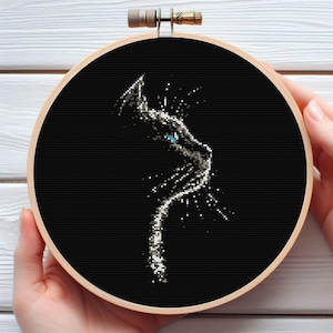 Puede incluir: Un bastidor de bordado de punto de cruz negro con un perfil de gato estilizado. El ojo del gato es azul brillante y el diseño está sobre un fondo negro. El bastidor de madera está sostenido por manos.