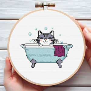 May include: A gray and white cat with closed eyes is taking a bath in a blue bathtub. The cat is surrounded by white bubbles. The bathtub has a purple towel hanging over the side.