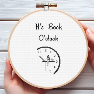 May include: A white embroidery hoop with a black and white design of a clock with a person sitting on the hand at the 12 o'clock position reading a book. The text "It's Book O'clock" is above the clock.