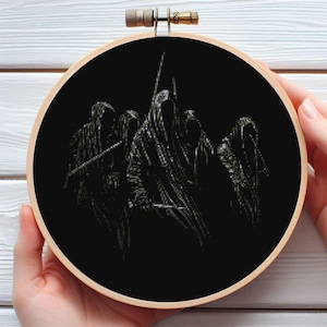 Può includere: Un telaio da ricamo a punto croce nero con un'immagine scura di figure incappucciate con armi. L'opera d'arte è ambientata su uno sfondo nero, incorniciata da un cerchio di legno. Il pezzo è tenuto dalle mani di una persona.