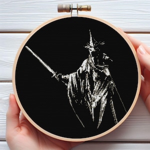 Puede incluir: Bordado de punto de cruz en blanco y negro en un aro de madera. El diseño presenta una figura con una espada, que lleva una corona y túnicas, sobre un fondo negro. El bordado está sostenido por dos manos.