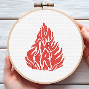 May include: A cross-stitch embroidery hoop featuring a red flame design with the word "FIRE" stitched within. The design is set on a white background and held in a wooden embroidery hoop.