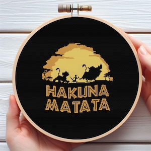 Puede incluir: Un diseño de punto de cruz en un aro de madera. El diseño presenta un fondo negro con una escena de puesta de sol dorada y las palabras "HAKUNA MATATA" en oro. La escena incluye siluetas de animales y árboles.
