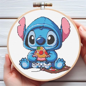 May include: A cross-stitch hoop featuring a blue cartoon character with large eyes and ears, wearing a hoodie and holding a slice of pizza. The character is sitting and holding a video game controller. The hoop is held by hands.