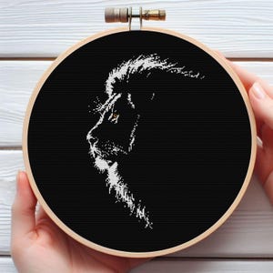 May include: A black and white cross-stitch lion portrait in a round wooden embroidery hoop. The lion's profile is depicted with white thread against a black background. The hoop is held by hands.