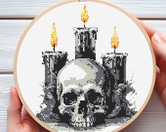 May include: Cross-stitch embroidery in a wooden hoop featuring a skull with three lit candles. The candles are gray with yellow and orange flames. The skull is gray and white, and the background is white.