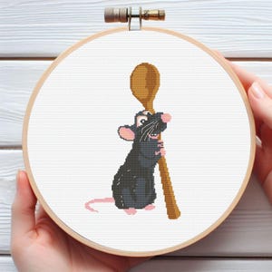 May include: Cross-stitch embroidery in a wooden hoop features a cartoon rat holding a wooden spoon. The rat is black with pink ears, feet, and tail. The spoon is brown, and the background is white.