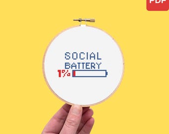 May include: A white cross-stitch hoop with the words "SOCIAL BATTERY" and a battery icon showing 1% charge. The text and icon are stitched in blue and red thread. The hoop is held by a hand against a yellow background.