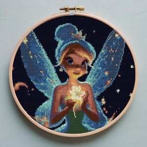 May include: A cross-stitch embroidery of a fairy in a wooden hoop. The fairy has blue wings and hair, a green dress, and is holding a glowing flower. The background is dark blue with scattered stars and sparkles.