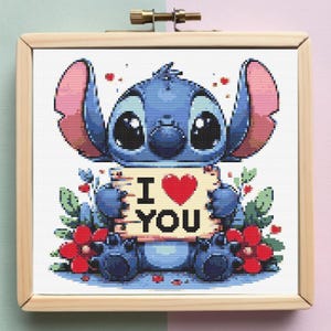 Puede incluir: Punto de cruz de Stitch sosteniendo un cartel que dice "I ❤️ YOU". El personaje azul está rodeado de flores rojas y hojas verdes, enmarcado en un bastidor de bordado de madera. Una obra de arte encantadora.