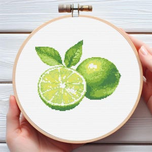 May include: Cross-stitch embroidery of two green limes, one sliced in half, with green leaves. The design is set in a wooden embroidery hoop. The embroidery is held by a person's hands.
