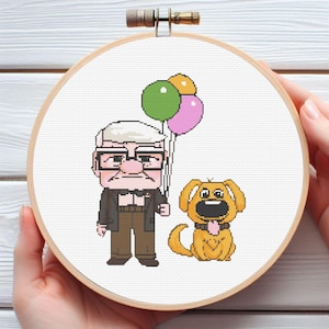 以下が含まれることがあります： 木製のフレームに入ったクロスステッチ刺繍。メガネをかけたおじいさんが風船と犬を持っている漫画です。男性はスーツと茶色のズボンを着用しています。犬はタン色で、黒い首輪をしています。風船は緑、ピンク、黄色です。