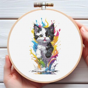May include: Cross-stitch embroidery of a black and white kitten with colorful paint splashes. The kitten is in a wooden embroidery hoop. The artwork features a playful design with vibrant colors, suitable for home decor.
