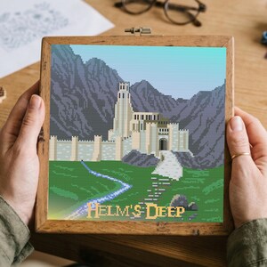 May include: A square cross-stitch embroidery in a wooden frame depicting a light-colored stone fortress nestled against dark purple mountains. A green field with a winding blue stream and a stone path leads to the castle gate. The text HELM'S DEEP is printed in yellow at the bottom.