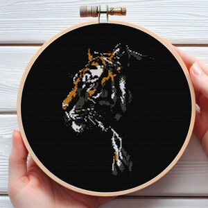 May include: A cross-stitch embroidery of a tiger's head in a wooden hoop. The tiger is depicted in shades of black, white, and orange against a black background. The embroidery hoop is held by a person.