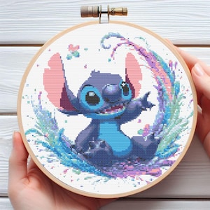 Puede incluir: Bordado de punto de cruz del personaje Stitch de Lilo & Stitch. La obra presenta a Stitch en tonos azules, con acentos rosas, rodeado de salpicaduras coloridas y mariposas, en un bastidor de bordado de madera.