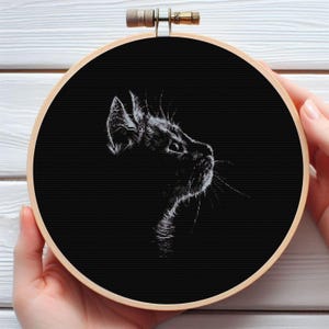Peut inclure: Broderie au point de croix en noir et blanc représentant le profil d'un chat. Le chat est brodé en fil blanc sur fond noir. Le cercle à broder en bois est tenu par des mains.