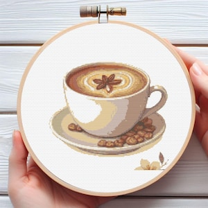 Pode incluir: Bordado em ponto cruz de uma xícara de café com um desenho de estrela de anis, sobre um pires com grãos de café. O bordado está em um bastidor de madeira, segurado por mãos. A paleta de cores inclui tons de marrom, bege e branco.