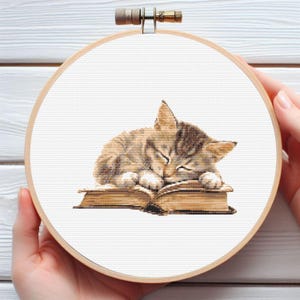 Pode incluir: Bordado em ponto cruz de um gatinho tigrado dormindo em um livro aberto. O gatinho é marrom e bege, com os olhos fechados. O livro é marrom e bege. O bordado está em um bastidor de madeira.