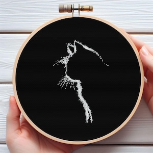 May include: A black cross-stitch embroidery hoop with a white cat silhouette. The cat's profile is detailed against the black fabric. The wooden hoop is held in place by a metal clasp.
