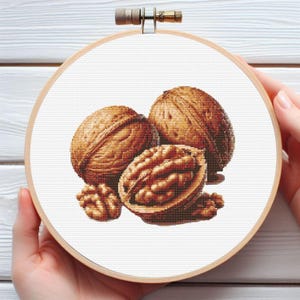 May include: A cross-stitch embroidery hoop featuring a detailed depiction of walnuts. The design includes whole and halved walnuts in shades of brown, set against a white background. The wooden hoop is held by a person's hands.