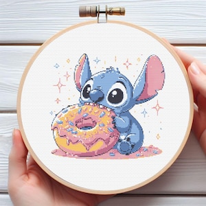 May include: A cross-stitch embroidery hoop featuring a blue cartoon character eating a pink frosted donut. The character has large eyes and large ears. The background is white with pink and yellow star accents.
