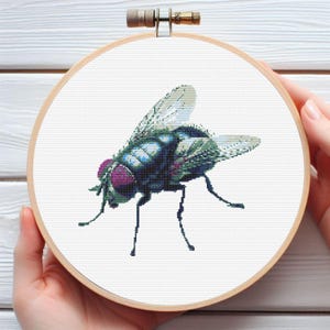 May include: A cross-stitch embroidery of a fly, featuring a detailed design with shades of blue, green, and purple. The fly is centered on a white fabric, framed by a wooden embroidery hoop. The artwork is held by hands.