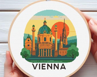 May include: A cross-stitch embroidery hoop featuring a pixelated cityscape of Vienna, Austria, with a teal and yellow sunset backdrop. The word "VIENNA" is stitched in black at the bottom. The hoop is held by hands.