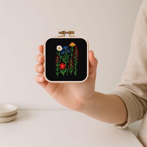 May include: A small, square cross-stitch embroidery hoop featuring a colorful floral design on a black background. The flowers include white daisies, blue cornflowers, red poppies, and yellow blooms. The hoop is held in a hand.