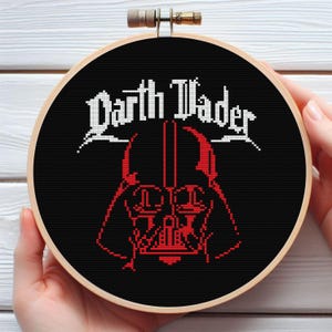 May include: A cross-stitch embroidery hoop featuring a black background with the words "Darth Vader" in white and a red Darth Vader helmet design. The embroidery is set in a wooden hoop.