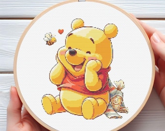 Puede incluir: Un bastidor de bordado de punto de cruz con Winnie the Pooh. El oso amarillo sonríe, lleva una camisa roja y una bufanda. Una abeja y un corazón también están cosidos en el diseño. El bordado está en un aro de madera.
