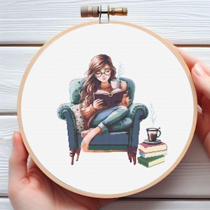 May include: A woman wearing glasses sits in a blue armchair reading a book and holding a cup of coffee. She is wearing a blue sweater and jeans. There are books stacked on a table next to her.