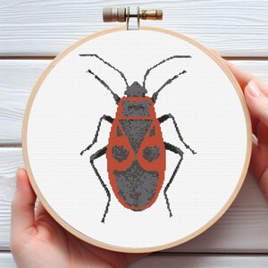 May include: Cross-stitch embroidery of a red and gray insect, possibly a firebug, on white fabric. The design is set in a wooden embroidery hoop. The insect has a detailed pattern and is centered on the fabric.