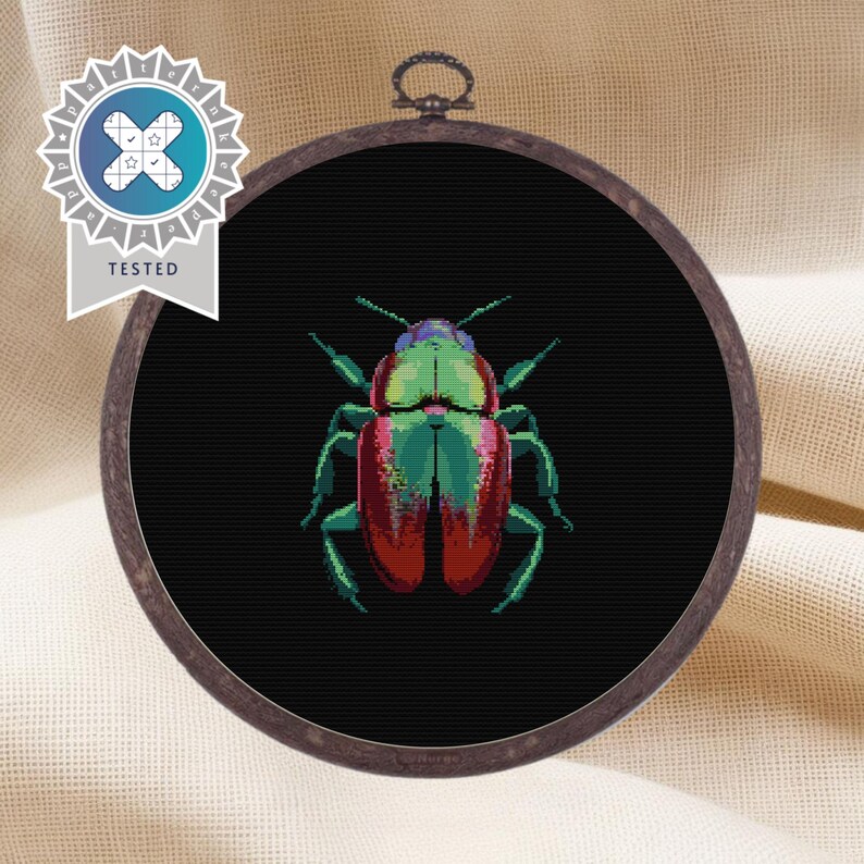 May include: A round embroidery hoop with a black background and a colorful beetle design. The beetle is green, red, and black with a metallic sheen.