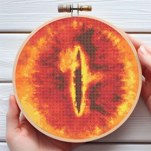 May include: A cross-stitch embroidery hoop featuring a fiery eye design in shades of red, orange, and yellow. The eye has a vertical pupil and is set against a dark background. The embroidery is framed in a wooden hoop.