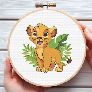 May include: A cross-stitch embroidery hoop depicting a cartoon lion cub with a smiling expression. The lion is in shades of yellow and brown, set against a white background with green foliage. The hoop is made of wood.