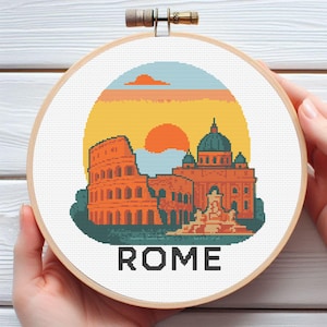 May include: Cross-stitch embroidery of Rome's Colosseum and a domed building, set against a sunset in orange, yellow, and blue. The word "ROME" is stitched in black at the bottom. The embroidery is displayed in a wooden hoop.