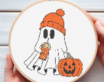 May include: A cross-stitch embroidery hoop featuring a Halloween-themed design. The design includes a ghost wearing an orange beanie and holding a drink, with a carved pumpkin beside it. The artwork is set against a white background.