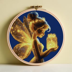 May include: A cross-stitch embroidery of a fairy holding a glowing jar, set against a dark blue background. The fairy is depicted in shades of gold and yellow, with detailed wings and hair. The artwork is framed in a round wooden hoop.