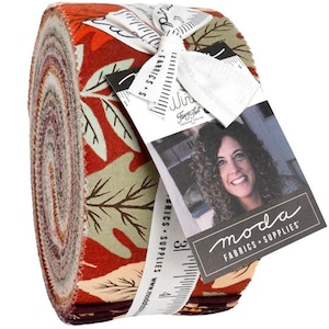 May include: A jelly roll fabric bundle featuring a repeating pattern of autumn-colored leaves in shades of burnt orange, cream, and dark brown. The roll is secured with a white ribbon printed with a ruler and a tag featuring a portrait and the text moda.